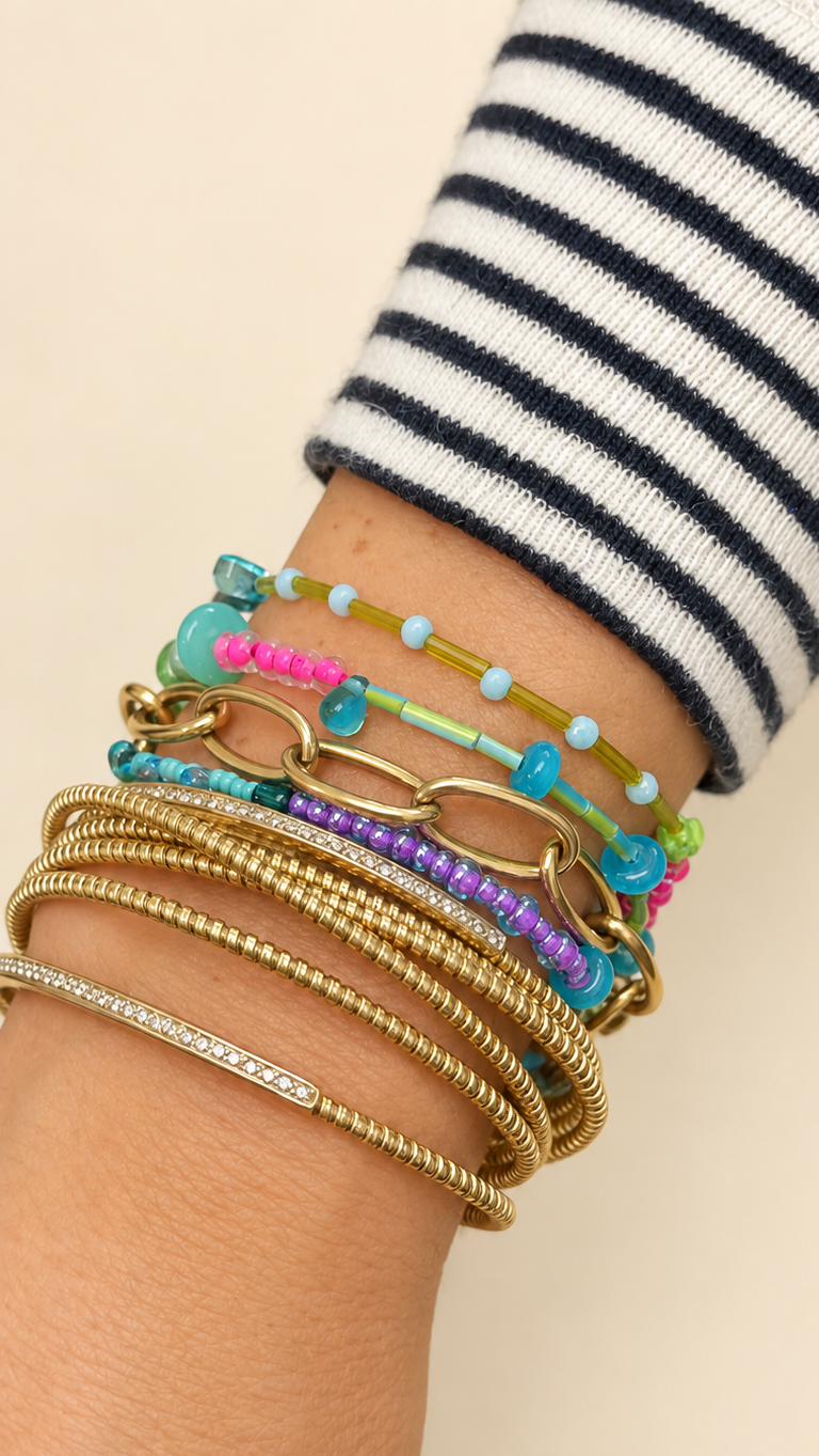 A close-up of a person's wrist wearing a stack of colorful beaded and metallic bracelets, including turquoise, pink, purple, and gold-tone chains, against a light beige background.
